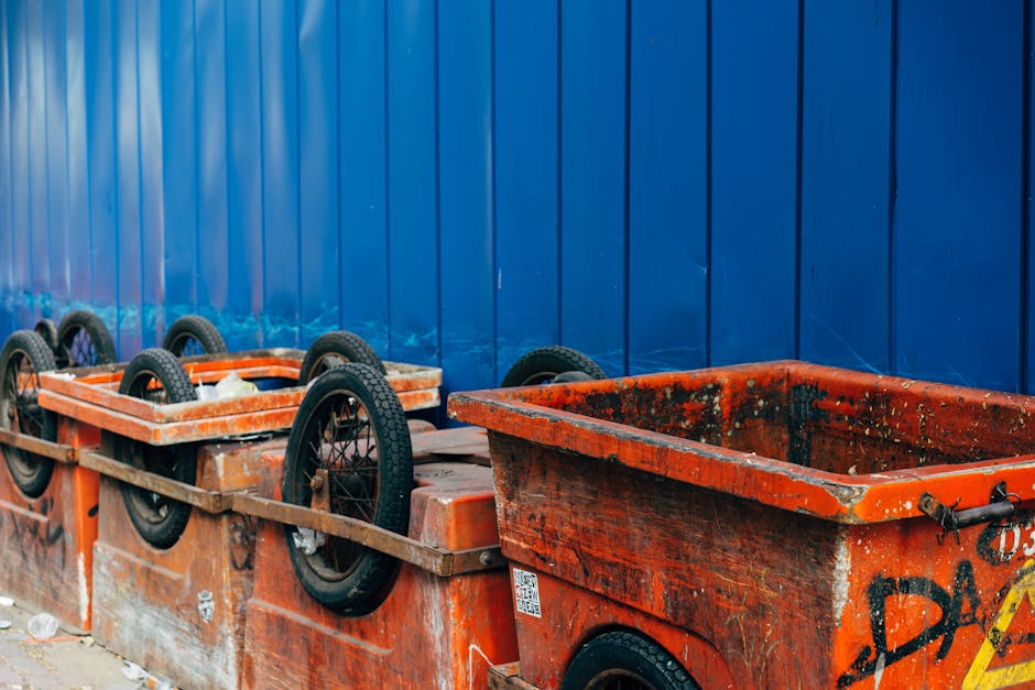 A row of overturned orange collection skip bins with black rubber tires visible on the sides, positioned on a paved surface against a tall blue metal fence with vertical panels. The bins are weathered, showing signs of rust, dirt, and paint chipping, with some graffiti and faded markings on their surfaces. One bin in the foreground has a partially open top, revealing residual debris and grime inside, while the others are closed or tipped over. The background features the blue fencing, suggesting an outdoor waste collection or storage area that may be used for various types of rubbish disposal or private waste handling, commonly associated with independent rubbish removal services. The scene is lit with natural daylight, casting minimal shadows, emphasizing the utilitarian environment typical for waste management operations.