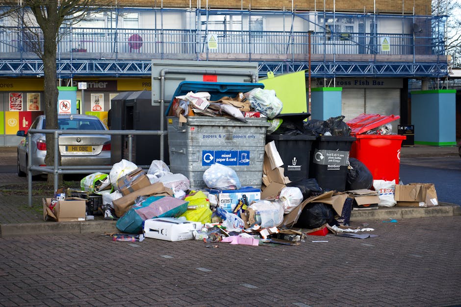 A large pile of mixed waste and rubbish bags is overflowing from several different waste containers situated on a paved area in front of a commercial building. The waste includes plastic bags, cardboard boxes, paper, and miscellaneous refuse, some spilling onto the surrounding pavement. The containers are black, blue, and red, with the blue bin marked for mixed paper and card, and the black one designated for general waste. The scene is outdoors, with a tree on the left side and a silver car parked nearby behind a metal railing. In the background, the building has a blue scaffolding structure covering the upper floor and storefronts beneath, one with a sign partially visible. The overall environment suggests an area designated for waste collection, possibly related to private rubbish removal services like those provided by Rubbish Removal Paddington, illustrating an example of bulk rubbish accumulation and the need for proper waste disposal or alternative rubbish collection options.