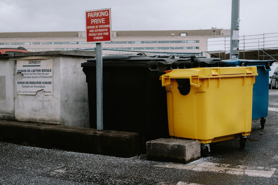 A row of overturned orange collection skip bins with black rubber tires visible on the sides, positioned on a paved surface against a tall blue metal fence with vertical panels. The bins are weathered, showing signs of rust, dirt, and paint chipping, with some graffiti and faded markings on their surfaces. One bin in the foreground has a partially open top, revealing residual debris and grime inside, while the others are closed or tipped over. The background features the blue fencing, suggesting an outdoor waste collection or storage area that may be used for various types of rubbish disposal or private waste handling, commonly associated with independent rubbish removal services. The scene is lit with natural daylight, casting minimal shadows, emphasizing the utilitarian environment typical for waste management operations.