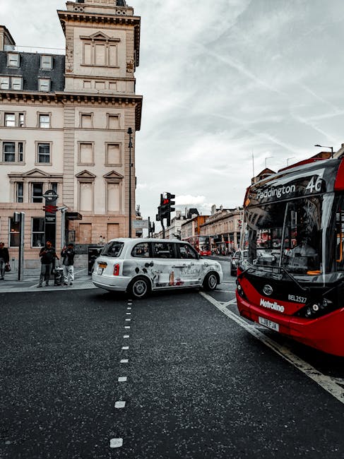 A busy urban street scene during daytime shows a white London taxi positioned near a pedestrian crossing, with the taxi’s side advertisement for an online delivery service partially visible. To the right, a red and black public bus displaying the route number 46 and destination Paddington is stopped, blocking part of the view. The background reveals a historic multi-storey building with ornate architectural details, light-colored stone façade, and multiple windows, along with other storefronts and street fixtures. Pedestrians are seen waiting at the crossing, some engaging in conversation or walking along the pavement. The sky is overcast with a light grey tone, adding a muted atmosphere to the scene. The environment appears to be a city centre intersection, consistent with locations where private and public transportation options such as taxis and buses are common, supporting the context of alternative methods to typical rubbish collection by illustrating typical city street activity.