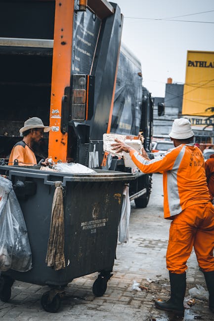 Two sanitation workers dressed in high-visibility orange uniforms and white hats are engaged in waste collection. One worker is standing on the pavement, holding a piece of rubbish and disposing of it into a large black wheeled bin attached to a waste collection truck. The bin has a textured surface with visible scratches and is positioned in front of the truck, which features a large, dark, and reflective rear section with orange safety markings. The background includes a yellow sign with black lettering, likely part of a commercial building, and partly visible structures or signage indicating an urban environment. Natural daylight illumination highlights the textures of the workers' uniforms, the metal surfaces of the waste truck, and the pavement, emphasizing the process of private waste handling typical of independent rubbish removal services like Rubbish Removal Paddington, ensuring a clear visual understanding of onsite rubbish collection in a city setting.