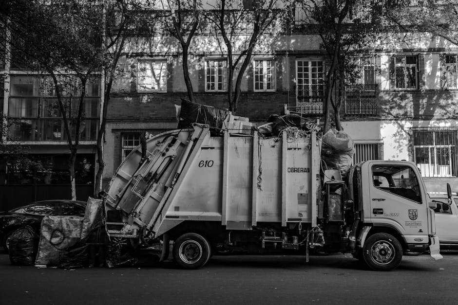 A black and white photograph depicts a waste collection truck parked on a city street with residential buildings in the background. The truck is a medium-sized commercial vehicle with a cab featuring a side mirror, and a large, enclosed rear compartment designed for collecting rubbish. The rear section of the truck is loaded with various waste materials, including large black plastic bags, cardboard boxes, and other debris, some of which are stacked or leaning against the sides of the truck. The waste appears to consist of mixed refuse, suitable for private rubbish collection services. Surrounding the vehicle are additional black plastic bags placed on the pavement, indicating active waste disposal or collection efforts. The background shows multi-story brick residential buildings with numerous windows and balconies, partially obscured by leafless trees casting shadows on the façade, suggesting an urban environment during the colder months. The overall scene is consistent with an alternative, on-site waste removal process facilitated by private services like Rubbish Removal Paddington, offering a practical solution for estate waste management in dense residential areas.