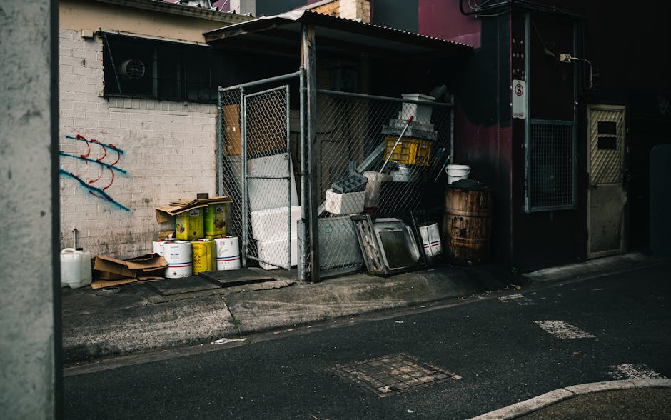 A narrow urban alleyway with a small outdoor storage area enclosed by a metal chain-link fence, situated against a white brick wall with a painted red and blue graffiti design. Inside the enclosure, there are various waste items, including large white plastic containers, metal trays, cardboard boxes, and yellow buckets stacked and leaning against the fence. On the concrete ground outside the enclosure, additional recycling containers or waste bins are visible along with a white plastic jug. To the right, a dark building wall features a small window with a black shutter and a door with a metal grate, adjacent to a wooden barrel and white bowl. The scene is lit with diffuse natural light, highlighting the clutter and materials associated with private waste disposal, reflecting typical rubbish accumulation in an urban setting, and illustrating a spot where independent rubbish removal services like those from Rubbish Removal Paddington might operate to clear such waste from private properties or alleyways.