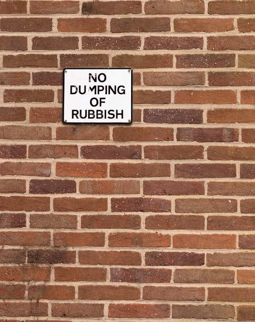A white rectangular sign with black text reading 'NO DUMPING OF RUBBISH' is mounted on a red brick wall. The bricks are arranged in a typical horizontal pattern with mortar joints in between, displaying variations in reddish-brown and dark brown tones. The sign is positioned approximately in the upper left quadrant of the wall, appearing to be securely affixed. The brick wall appears clean and well-maintained, with no visible graffiti or damage, serving as a backdrop for the sign that warns against illegal dumping. The lighting is even, suggesting natural daylight, and there are no other objects or environmental features visible in the scene. This setting subtly relates to waste management and the importance of adhering to local rubbish disposal regulations, aligning with the context of private waste handling services such as those offered by Rubbish Removal Paddington.