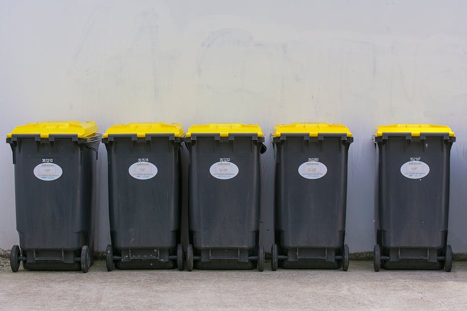 A narrow urban alleyway with a small outdoor storage area enclosed by a metal chain-link fence, situated against a white brick wall with a painted red and blue graffiti design. Inside the enclosure, there are various waste items, including large white plastic containers, metal trays, cardboard boxes, and yellow buckets stacked and leaning against the fence. On the concrete ground outside the enclosure, additional recycling containers or waste bins are visible along with a white plastic jug. To the right, a dark building wall features a small window with a black shutter and a door with a metal grate, adjacent to a wooden barrel and white bowl. The scene is lit with diffuse natural light, highlighting the clutter and materials associated with private waste disposal, reflecting typical rubbish accumulation in an urban setting, and illustrating a spot where independent rubbish removal services like those from Rubbish Removal Paddington might operate to clear such waste from private properties or alleyways.