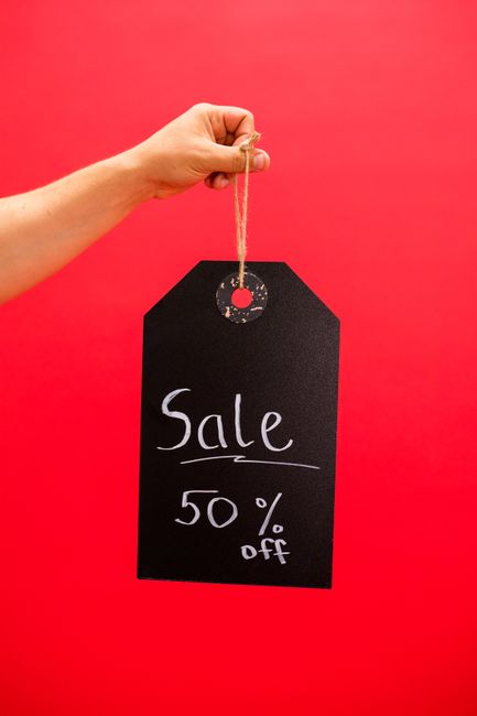 A light-skinned hand holds a small string attached to a black rectangular sale tag with rounded top corners against a vibrant red background. The tag features handwritten white chalk or marker text that reads 'Sale 50% off'. The background is uniform and bright, drawing attention to the tag and hand, with no other objects or environmental details visible. The scene's focus is on promoting a discount, and the image style is minimalistic, emphasizing the sale message in a clean and straightforward manner, suitable for retail or promotional contexts related to discount offers.