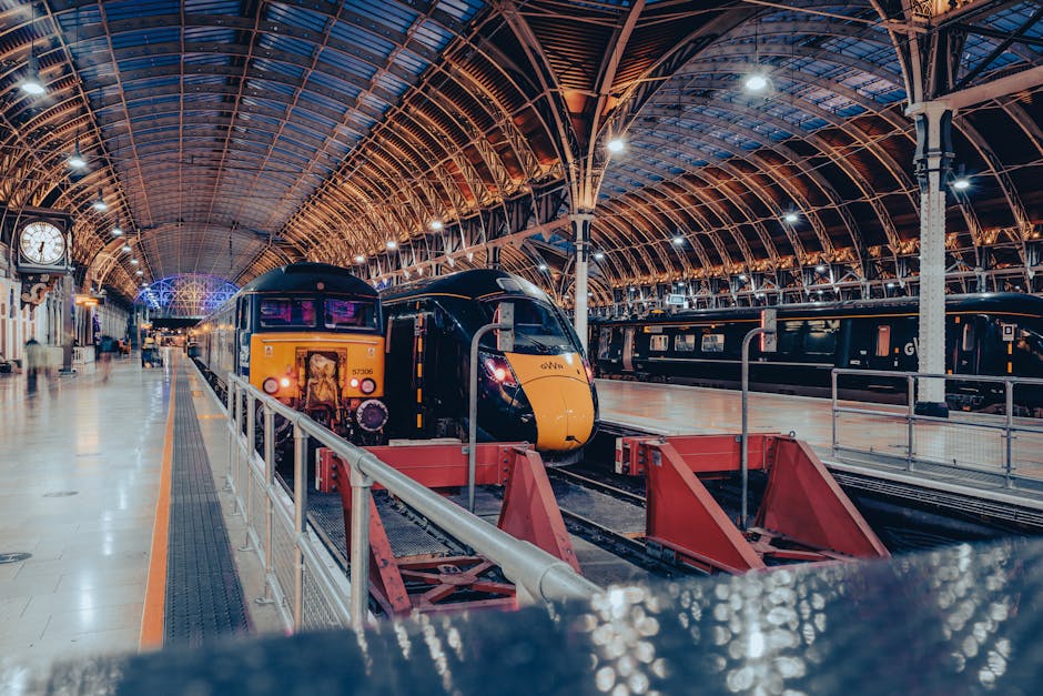 A modern high-speed train with a sleek black and yellow exterior is stationed inside a spacious, historic railway station with an arched, metal and glass roof illuminated by bright overhead lighting. The train's front end features a smooth, aerodynamic design, and it is positioned on tracks that are separated by red safety barriers. To the right, there are additional train carriages with dark, polished finishes, parked along adjacent platforms. On the left side of the image, a clock mounted on the wall displays the time, and the station's interior includes tiled flooring, metal railings, and a few illuminated signs, all contributing to a clean and well-maintained environment. The scene is quiet, with no visible passengers or personnel, emphasizing the station's orderly, ready-for-service atmosphere. The lighting reflects off the train’s shiny surface, highlighting its modern design, while the station's architecture, with its intricate roof framing, adds a historic touch to the setting, subtly illustrating the contrast between traditional railway design and contemporary transport technology, aligning with the context of efficient, private rail-based or alternative transport solutions in urban environments. Rubbish Removal Paddington occasionally provides independent waste handling services for station facilities, supporting clean and organized public spaces.