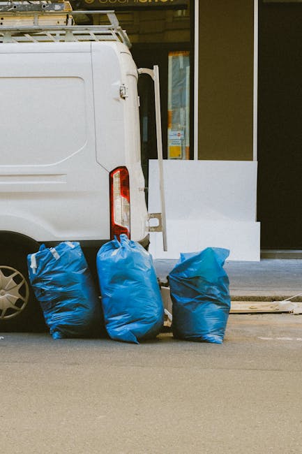 A white delivery van parked on a street near a building with large glass windows and a yellow frame. The rear of the van is visible, with three large blue rubbish bags placed on the pavement close to the back door. The bags appear to be filled with waste and are made of plastic with a slightly crinkled texture. Behind the bags, a white panel or barrier is positioned near the building's entrance, which has a clear glass door. The environment suggests a commercial or residential area, and the scene indicates a private rubbish collection, typical of independent waste handling services such as Rubbish Removal Paddington, operating in the local vicinity. The lighting is natural, likely indicating daytime, with minimal shadows, and the street surface is smooth asphalt. Overall, this image depicts a typical scene of rubbish awaiting collection, highlighting the presence of external waste disposal solutions outside a modern storefront or property.