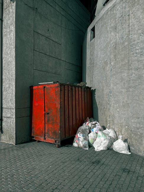 A red metal waste dumpster with visible rust and chipped paint is positioned in a narrow outdoor space, situated against a dark green wall on the left and a light grey concrete wall on the right. Several clear plastic bags filled with mixed rubbish, including paper, plastic bottles, and packaging, are piled up beside the dumpster on the paved ground, which consists of dark grey interlocking bricks. The environment appears to be an alleyway or service area adjacent to a building, with minimal natural lighting and clean, sturdy surface materials that support off-site or private waste collection activities, characteristic of independent rubbish removal services. The scene reflects an area designated for temporary rubbish storage awaiting collection or disposal, aligning with the context of flexible waste handling mechanisms in urban settings.