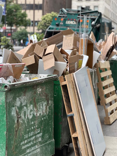 A large green rubbish skip filled with assorted cardboard boxes, some flattened and others intact, along with a few white foam containers and pieces of packaging. The cardboard varies in shades of brown, with some packaging bearing labels or printed markings. The heap of waste extends beyond the edges of the skip, which is placed on an urban street. To the right, there is a wooden pallet leaning against the side of the skip, partially obscuring the view. In the background, a large green waste collection truck with branding visible is parked, with its rear end elevated for loading. Surrounding the scene are trees, buildings, and parked vehicles, indicating a city environment. The image appears to be taken during daylight with natural light illuminating the sites, capturing a typical scenario of private or independent waste disposal involving rubbish removal services such as those offered by Rubbish Removal Paddington, focused on bulky waste collection or on-site clearance.