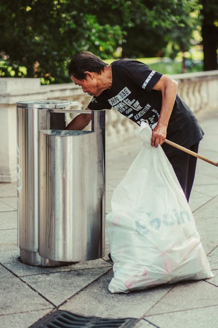 A middle-aged man with dark hair, wearing a black T-shirt with white printed text, is seen on a paved outdoor area disposing of waste into a cylindrical metal rubbish bin. He is bent slightly forward with his right arm extended into the open-top bin, which appears to be made of stainless steel and has a modern, reflective finish. In his left hand, he holds a large white plastic garbage bag that is partly filled with waste. The man is using a wooden stick or broom handle to assist in placing the rubbish into the bin. The background features a low stone or concrete wall with decorative balustrades and lush green trees, indicating a park or public walkway setting. The natural daylight and shadows suggest a bright, clear day. This scene exemplifies independent waste disposal, which may be an alternative to official rubbish collection services, aligning with private waste handling practices often managed by specialist firms like Rubbish Removal Paddington.