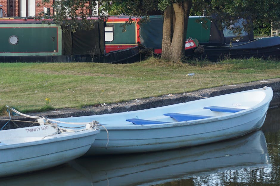 A white rowboat with three blue plastic seats is moored on a calm waterway in the foreground, with a slightly weathered surface showing minor dirt and rust marks. The boat is tethered with white ropes attached to a nearby structure. Behind the boat, there is a grassy embankment with short, patchy green grass and a few scattered yellow flowers, leading up to a row of narrowboats docked along the bank. The narrowboats are painted in dark green, black, and red colors, featuring rectangular windows, some with white frames, and black protective coverings on parts of their structures. A large tree with a thick, textured trunk and dense green foliage stands at the edge of the grass, partially obscuring the narrowboats. The scene is illuminated by natural daylight, creating clear reflections of the boats on the water’s surface, suggesting a quiet, residential or leisure environment. This setup reflects typical private waterfront properties and incidental waste removal considerations often seen near such residential water features, managed by local services like Rubbish Removal Paddington.