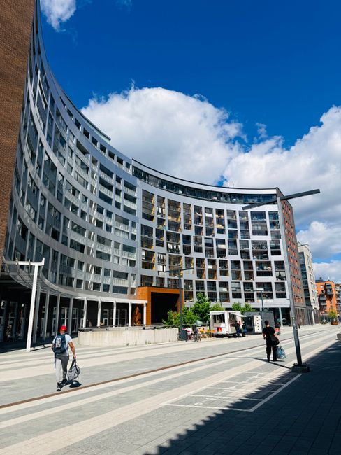A modern multi-storey building with a curved facade, featuring numerous rectangular windows with dark frames and some glass balconies, is set against a bright blue sky with scattered white clouds. The building's exterior combines white and grey tones, with a prominent orange entrance area at ground level. In the foreground, there is a wide paved pedestrian area with light and dark grey tiles, where two individuals are walking; one carrying a backpack and the other pulling a small suitcase or trolley bag. To the right, a streetlight and a small roadside vendor stand are visible, alongside some greenery including small bushes and young trees near the building entrance. The scene conveys a contemporary urban environment, which Rubbish Removal Paddington might serve by providing private waste disposal solutions for residential and commercial properties within the city, complementing traditional council waste collection through independent rubbish removal services.